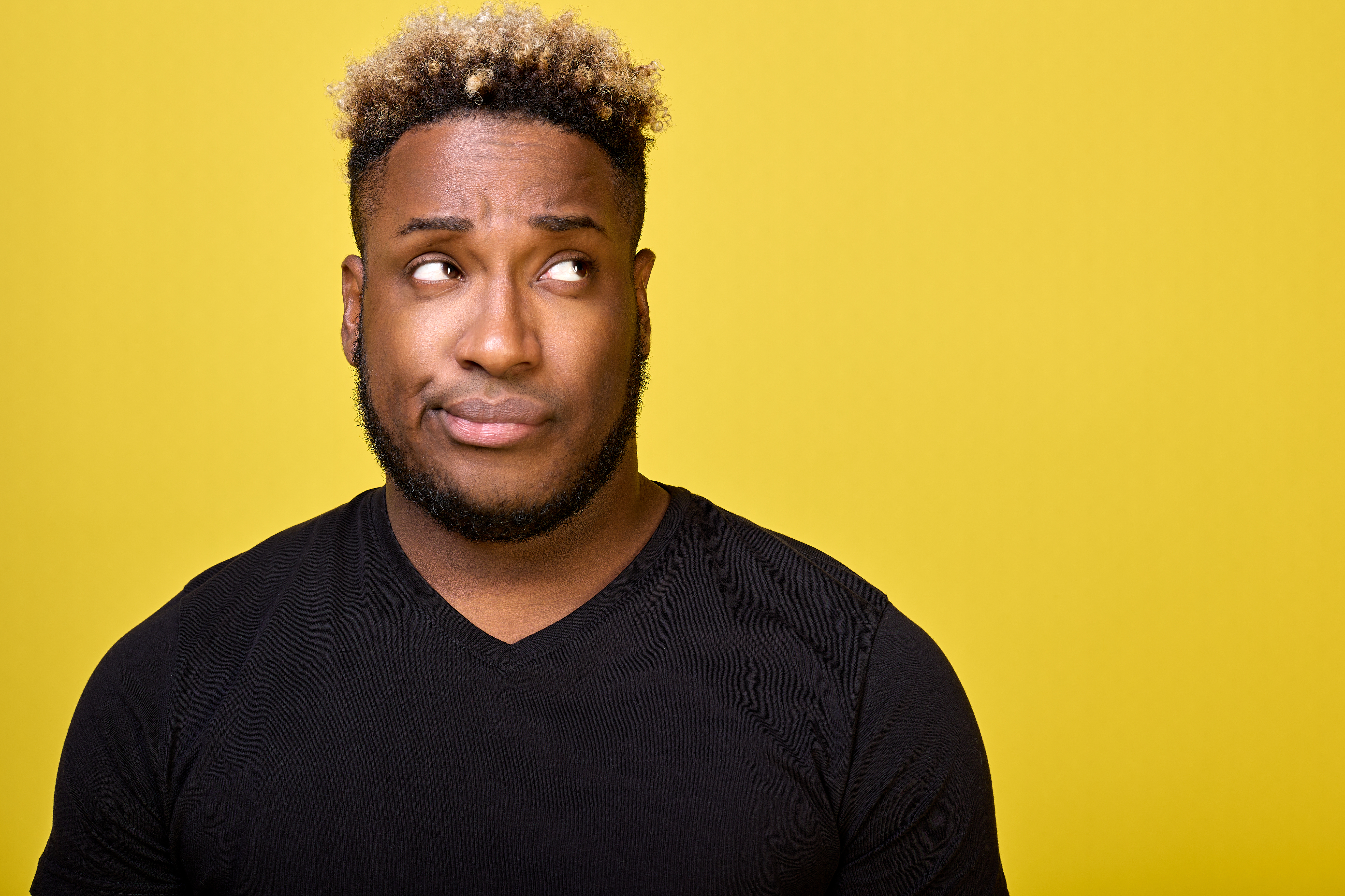 A dark-skinned man with a beard with a smirk on his face looks to the side in disapproval, faced with a difficult choice, dressed in an everyday black T-shirt. Doubting guy looks up with a smirk
